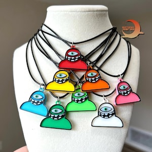 May include: A collection of colorful pendant necklaces featuring a cartoonish cyclops design. Each pendant is a different color, including red, yellow, orange, blue, green, pink, and white, with a black outline and a black cord.
