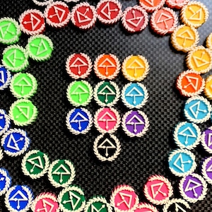 May include: A collection of colorful, circular game tokens. Each token features a white arrow and a stylized letter within a colored circle, surrounded by a white wreath. The tokens are arranged on a textured black surface.