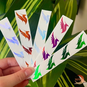 May include: A set of six iridescent vinyl stickers featuring a phoenix design in various colors, including red, purple, green, and blue. The stickers are on white sheets and are perfect for adding a touch of sparkle to any project.