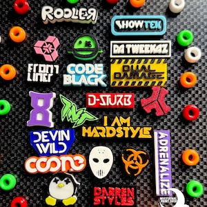 May include: A collection of colorful shoe charms and beads on a textured black surface. The charms feature various designs and text, including "ROOLER," "SHOWTEK," and "DA TWEEKAZ." The beads are in shades of red, orange, green, and white.