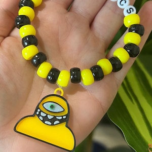 May include: A black and yellow beaded bracelet with a charm of a yellow cartoon character with one eye and a toothy grin. The charm is hanging from the bracelet.