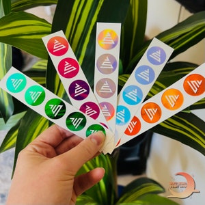 May include: A hand holding several sticker sheets with round, colorful stickers. Each sticker features a white geometric design inside a circle of various colors, including green, pink, purple, and orange. The stickers have a shiny, holographic finish.
