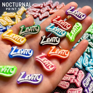 May include: A collection of small, colorful shoe charms featuring the word Levity in a stylized, raised font. These individual plastic charms come in various solid colors including red, blue, yellow, green, purple, orange, black, and pink.