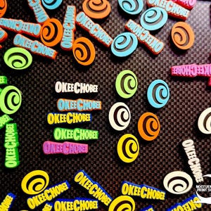 May include: A collection of colorful, decorative charms and embellishments. The charms feature the text "OKEE CHOBEE" in various colors, including green, orange, blue, and pink. Some charms have a swirl design. The background is a dark, textured surface.