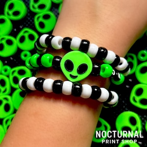May include: Three stacked bracelets featuring alternating black and white round beads. The center bracelet includes a bright neon green alien head charm with black eyes and a small mouth. The text Nocturnal Print Shop is visible at the bottom.