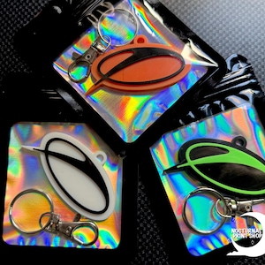 May include: Keychains in orange, white, and green with a black design, attached to silver keyrings. The keychains are packaged in clear, iridescent pouches. The background is a dark, textured surface.