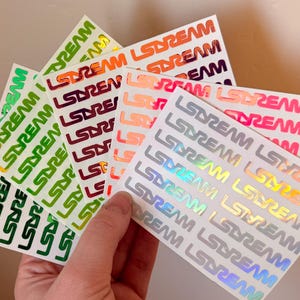 May include: A hand holds several rectangular stickers with the word "LSDREAM" printed repeatedly in various colors. The stickers feature green, orange, red, pink, and holographic silver text on a white background. The stickers are arranged in a fan shape.
