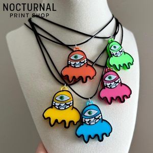 May include: Five necklaces featuring colorful, dripping ghost-shaped pendants on black cords. Each pendant has a single green eye and a wide, toothy grin. The colors include orange, lime green, pink, yellow, and blue. Text reads NOCTURNAL PRINT SHOP.