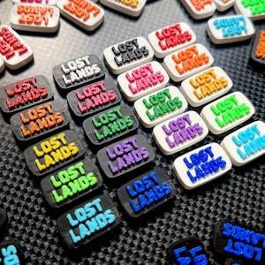 May include: A collection of rectangular shoe charms featuring the text LOST LANDS in raised lettering. The charms are available in various color combinations, including black, white, red, blue, green, orange, purple, and light blue, arranged in rows.