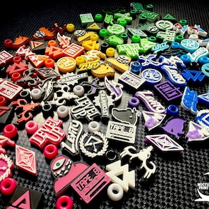 May include: A collection of colorful, decorative shoe charms with various designs and text, including "LEVEL UP", "TAPE B", and "GRIZ". The charms are in a variety of shapes and colors, including red, pink, yellow, green, blue, and purple.
