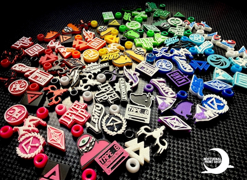 May include: A collection of colorful, decorative shoe charms with various designs and text, including "LEVEL UP", "TAPE B", and "GRIZ". The charms are in a variety of shapes and colors, including red, pink, yellow, green, blue, and purple.