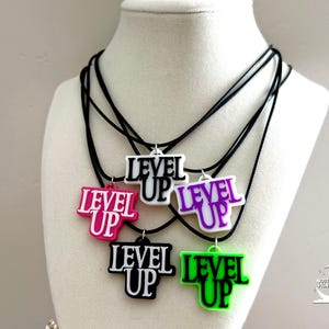 May include: A collection of "Level Up" necklaces in various colors, including pink, white, purple, black, and green. Each pendant features the words "Level Up" in a bold, contrasting font, suspended from a black cord.