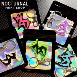 May include: Five keychains in black plastic pouches with holographic inserts. Each keychain features a jagged, stylized graphic design in pink, purple, white, black, or light blue. Each includes a silver metal split ring and a lobster clasp.