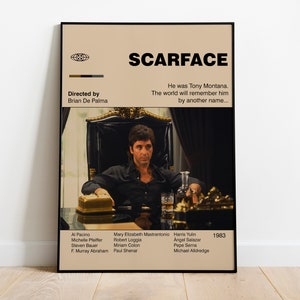 May include: A framed movie poster for "Scarface" featuring Al Pacino. The poster has a beige background with the movie title in bold black letters. The image shows a man in a black shirt seated in a chair. The poster includes the director's name and the year 1983.