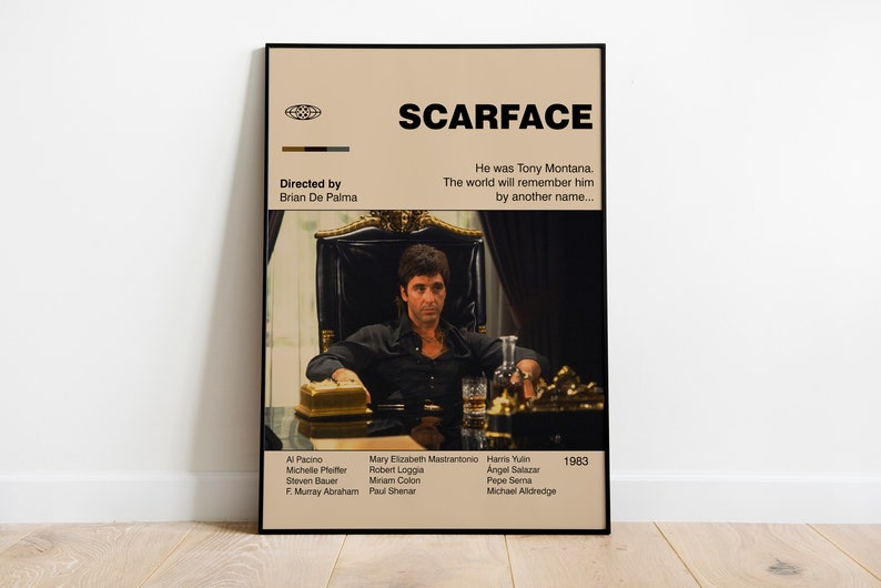 May include: A framed movie poster for "Scarface" featuring Al Pacino. The poster has a beige background with the movie title in bold black letters. The image shows a man in a black shirt seated in a chair. The poster includes the director's name and the year 1983.