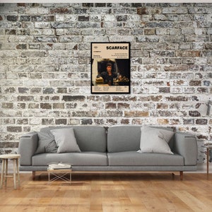 May include: A framed movie poster for "Scarface" hangs on a brick wall. The poster features a beige background with the movie title and a photo of the main character. The poster is in a black frame and is displayed above a gray sofa.
