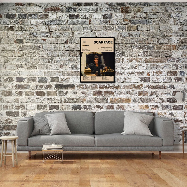 May include: A framed movie poster for "Scarface" hangs on a brick wall. The poster features a beige background with the movie title and a photo of the main character. The poster is in a black frame and is displayed above a gray sofa.