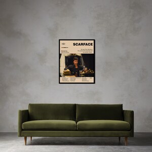 May include: A framed movie poster for "Scarface" featuring a seated man at a table with a drink and a hat. The poster has a beige background with black text and a black frame. The poster is displayed above a green sofa.