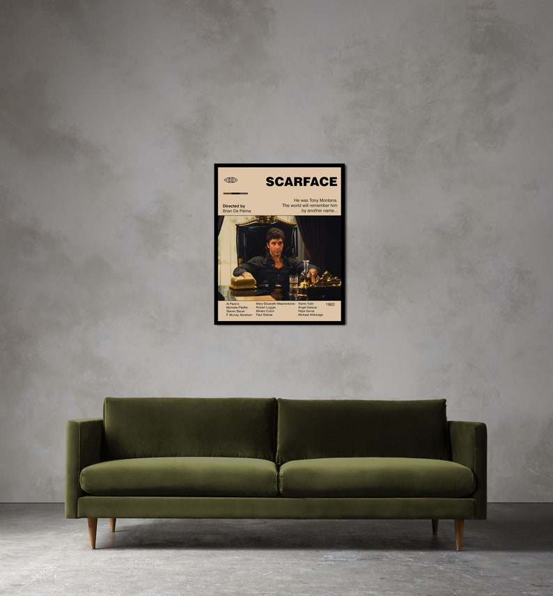 May include: A framed movie poster for "Scarface" featuring a seated man at a table with a drink and a hat. The poster has a beige background with black text and a black frame. The poster is displayed above a green sofa.