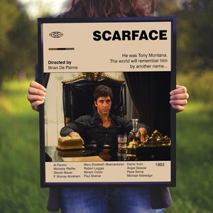 May include: A framed movie poster for "Scarface" featuring Al Pacino. The poster has a tan background with the movie title in bold black letters. The image shows Tony Montana seated at a table. The poster includes the director's name and the year 1983.