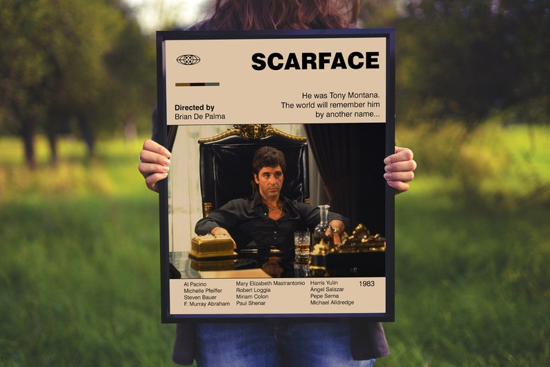 May include: A framed movie poster for "Scarface" featuring Al Pacino. The poster has a tan background with the movie title in bold black letters. The image shows Tony Montana seated at a table. The poster includes the director's name and the year 1983.