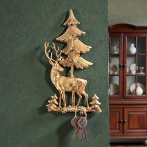 May include: A gold-colored metal key holder featuring a deer and pine tree design. The key holder is mounted on a dark green wall and holds a set of keys. The design is detailed and decorative.