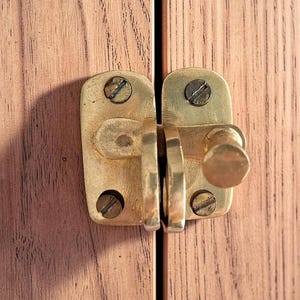 Puede incluir: Primer plano de un pestillo de armario de latón con pomo y tornillos, montado en una puerta de armario de madera marrón claro. El pestillo es rectangular con bordes redondeados y un pomo circular.