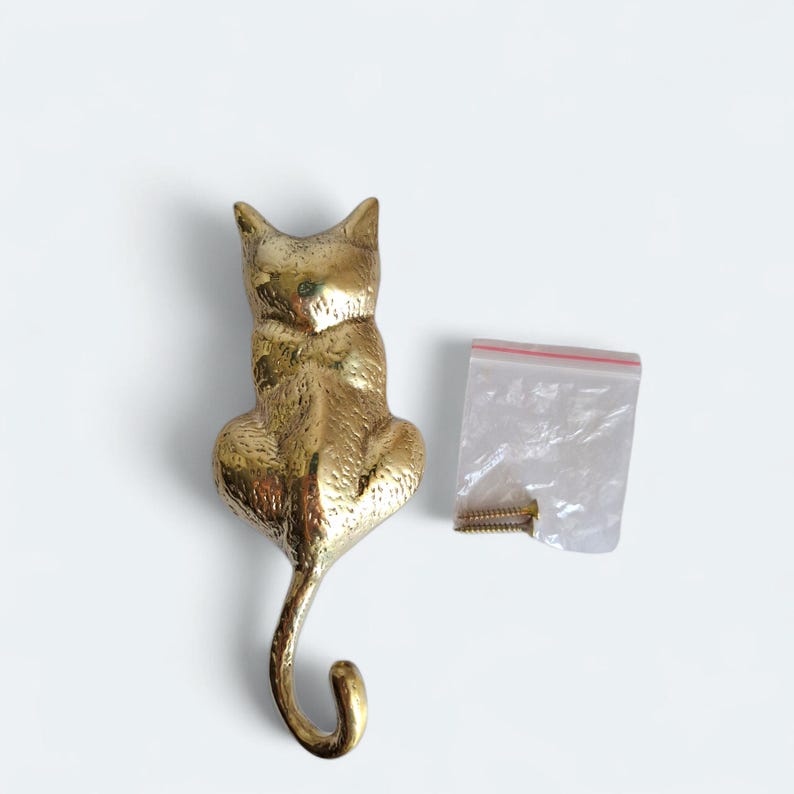 May include: A golden-colored cat-shaped wall hook. The hook is made of metal and has a curved tail forming the hook. A small plastic bag with screws is next to the hook. The cat is sitting upright.