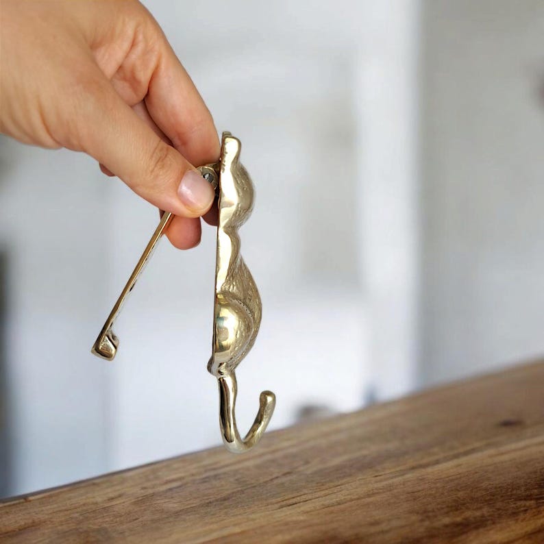 May include: A brass cat-shaped wall hook with a curved hook and a hinged arm. The hook is held by a hand, showcasing its polished finish and decorative design. The hook is designed to be mounted on a wall.