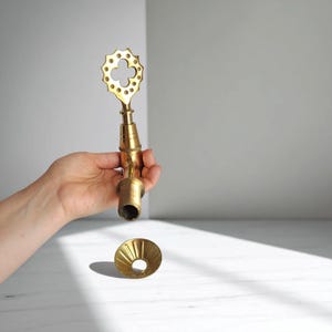 May include: A brass faucet with a decorative clover-shaped handle and a flared base. The faucet is held in a hand against a neutral background. The faucet is a warm gold color.