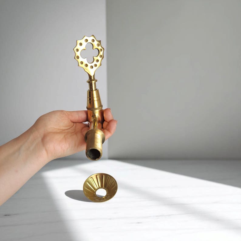 May include: A brass faucet with a decorative clover-shaped handle and a flared base. The faucet is held in a hand against a neutral background. The faucet is a warm gold color.