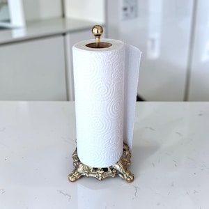 Può includere: Un portarotolo verticale con base in metallo color oro dotata di piedini decorativi ornati e un'asta centrale sormontata da un piccolo terminale sferico. È montato un rotolo di carta assorbente bianca con un motivo circolare in rilievo.