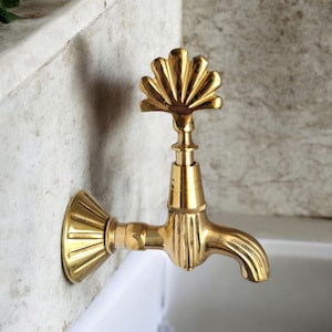 May include: A brass-coloured tap with a decorative fan-shaped handle. The tap is wall-mounted and has a curved spout. The design includes ribbed detailing and a flared base, suggesting a vintage or antique style.