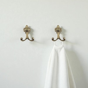May include: Two antique brass wall hooks with a decorative top and three curved hooks. One hook holds a white towel. The hooks are mounted on a white wall. Bathroom decor.