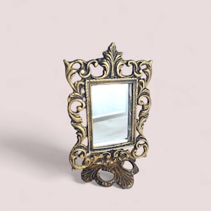 Peut inclure: Un petit miroir de courtoisie orné avec un verre rectangulaire. Le cadre présente un motif tourbillonnant détaillé en or et noir. Le miroir repose sur une base décorative, adapté à une coiffeuse ou un bureau.