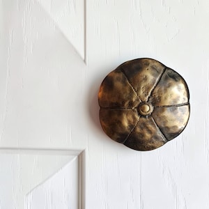 May include: A circular, bronze-toned metal door knob with a textured, hammered surface. The knob features a segmented, flower-like design with six distinct petals radiating from a small, smooth, raised circular center point.