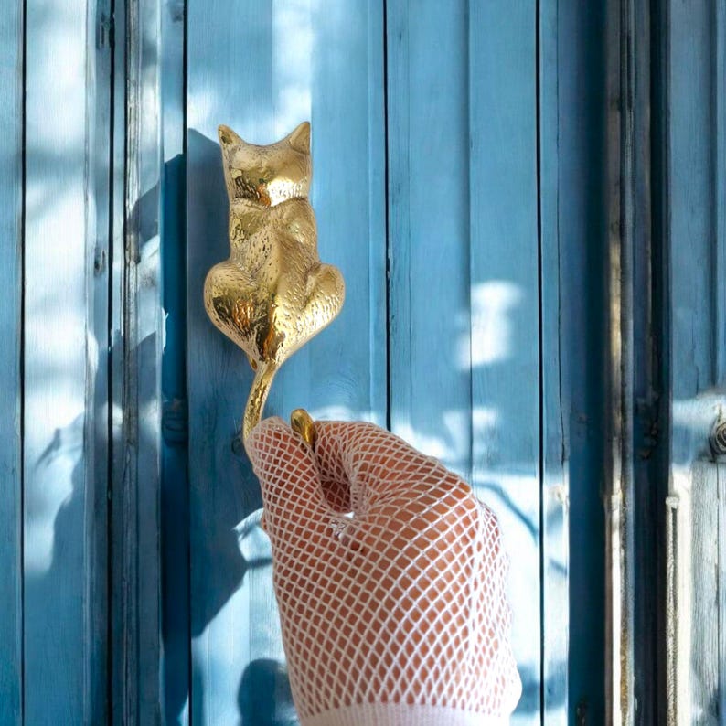 May include: A gold-colored cat-shaped door knocker mounted on a blue wooden door. The knocker is being held by a hand wearing a white fishnet glove. The door has vertical wooden panels.