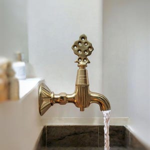 May include: A gold-toned faucet with an ornate design on the spout, pouring water into a gray stone sink.