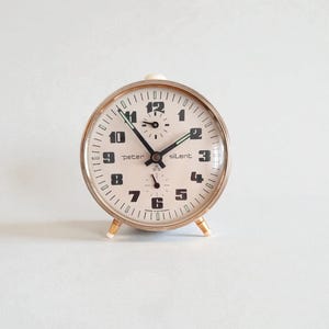 Könnte beinhalten: Ein Vintage-Wecker mit cremefarbenem Zifferblatt und silberfarbenem Metallrahmen. Die Uhr hat schwarze Ziffern, Zeiger und den Text "peter silent". Sie hat goldfarbene Füße und eine weiße Oberseite. Ein klassisches Design.