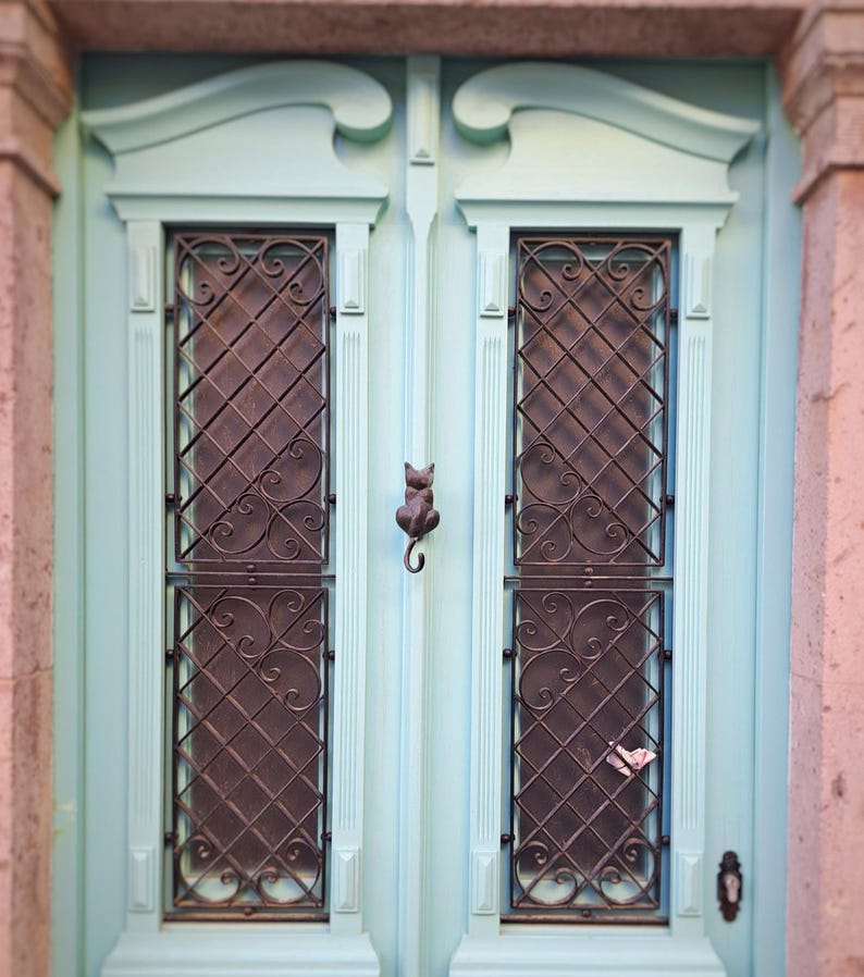 May include: A light blue wooden door with two rectangular windows featuring decorative iron grilles. A cat-shaped door knocker is centered on the door. The door is framed by pink stone columns.