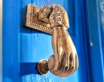 May include: A gold-toned door knocker shaped like a hand, with a detailed design. The knocker is mounted on a blue door.