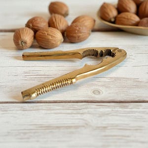 Pode incluir: Um quebra-nozes dourado com uma pega canelada e um mecanismo de quebra dentado. Várias nozes castanhas estão espalhadas à volta do quebra-nozes, com mais numa pequena tigela no fundo. O quebra-nozes está numa superfície de madeira clara.