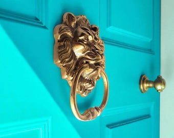 May include: A gold-colored lion head door knocker with a ring handle is mounted on a bright turquoise door. A matching gold door handle is visible to the right. The door has a paneled design.