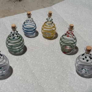 May include: Six decorative glass bottles with cork stoppers. Each bottle features a unique design with colored stripes and small decorative glass beads. The bottles are arranged on a white surface.
