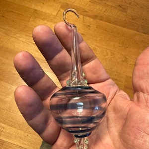Puede incluir: Adorno navideño de cristal transparente con un gancho. El adorno tiene un centro bulboso con rayas azules horizontales y una parte inferior puntiaguda. Pequeños detalles verdes son visibles cerca de la parte superior e inferior.