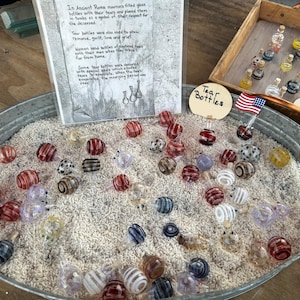 May include: A close-up of a metal container filled with small, colorful glass bottles. The bottles are arranged in a random pattern and are surrounded by white sand. A sign reads "Tear Bottles".