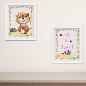 May include: Two framed watercolor prints and a knitted bunny on a wooden surface. One print features a beaver with a flower, the other says "Life is Sweet!" The bunny is beige with a pink tail.