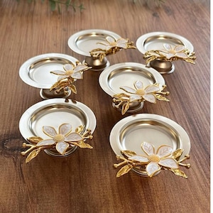 May include: A set of six silver-coloured metal coasters, each with a gold-coloured floral design. The coasters have a small, round dish on a pedestal base. The floral design features white petals and gold-coloured leaves.