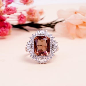 May include: A silver ring featuring a large, rectangular, brown gemstone surrounded by smaller, clear, baguette-cut stones. The ring's design is reminiscent of a halo, with the central stone as the focal point. The ring is set against a soft, neutral background.