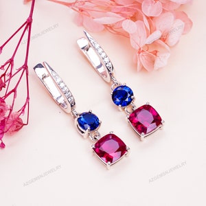 Może przedstawiać: Para srebrnych kolczyków wiszących z niebieskimi i czerwonymi kamieniami szlachetnymi. Kolczyki mają mały okrągły niebieski kamień szlachetny, większy kwadratowy czerwony kamień szlachetny i rząd małych przezroczystych kamieni szlachetnych na obręczy.